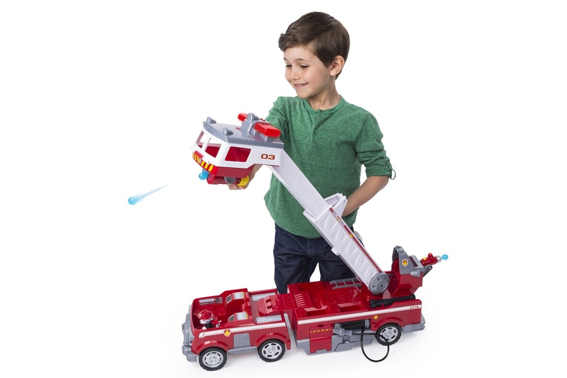 A Paw Patrol fire truck toy with a 2 ft. tall extendable ladder, mini fire cart, light and sounds, and water cannon launchers. A boy is kneeling behind the toy, exploring the features of the fire truck.