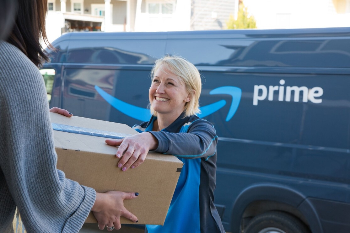 A woman delivers a box to another woman. The delivery driver stands in front of an Amazon DSP van. 